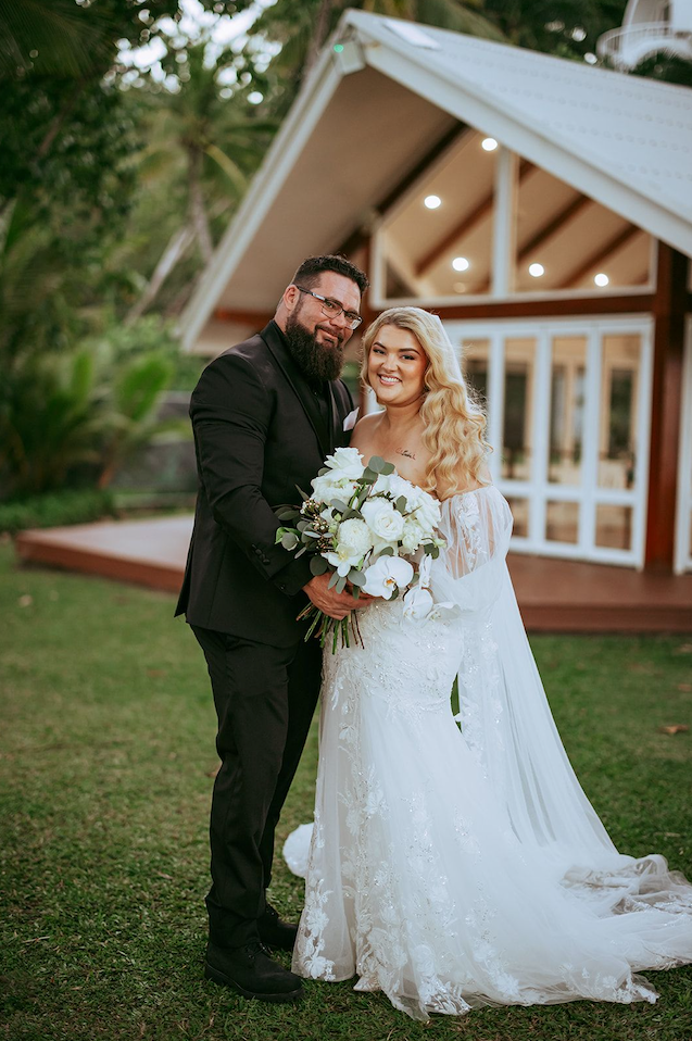 Newlyweds posing in front of a small chapel, bride in a white gown holding a bouquet, groom in a black suit