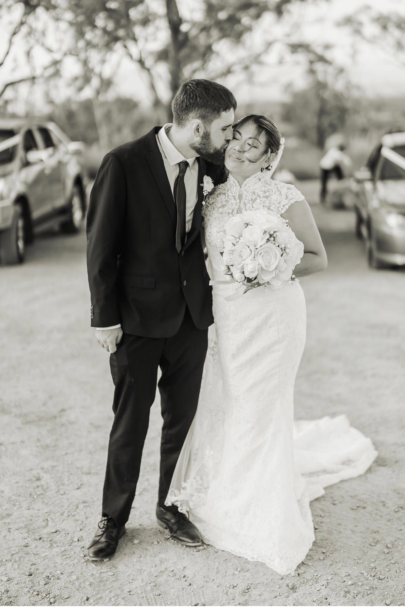 Wedding couple standing together outdoors, smiling and touching foreheads, with cars in the background
