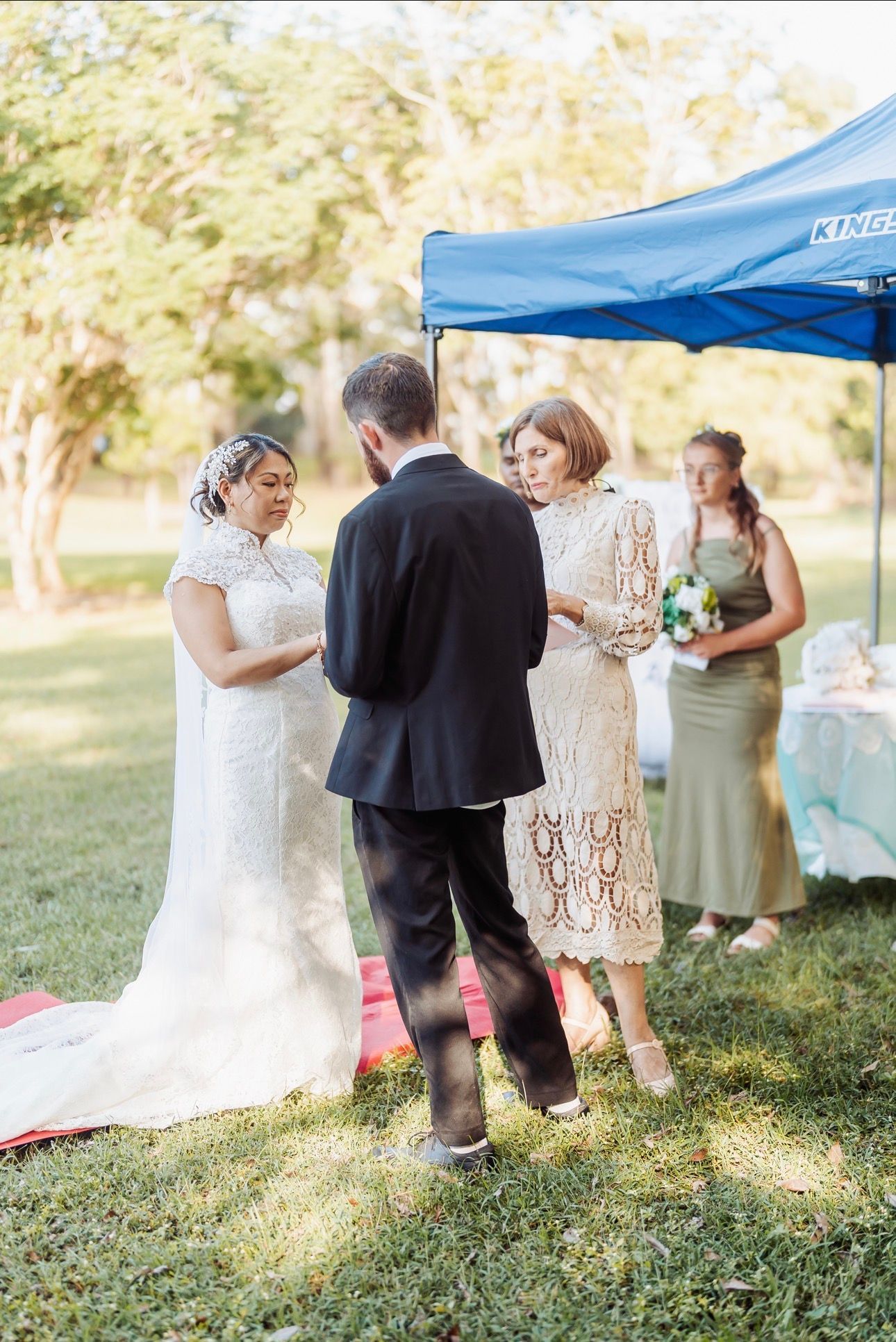 Outdoor wedding ceremony with bride, groom, and officiant under a blue canopy in a sunlit garden