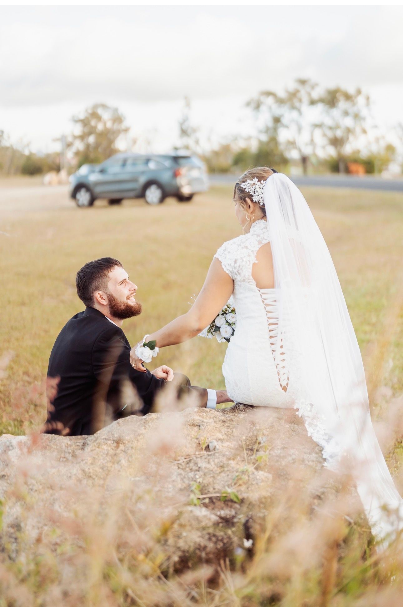 Bride helps groom kneel in a grassy field, with a blue SUV in the background.