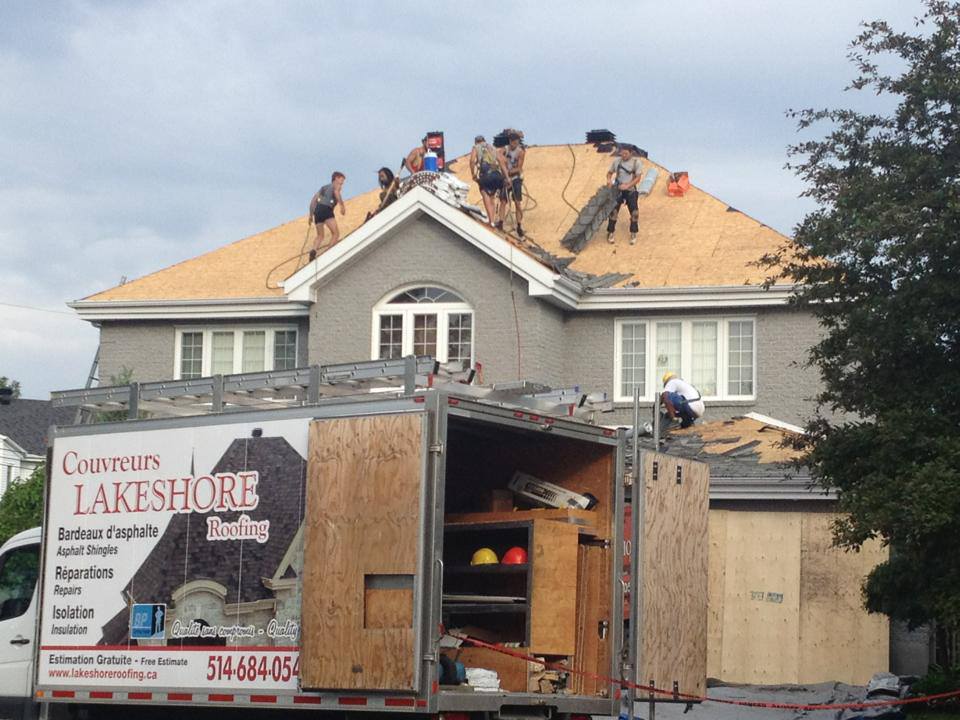 Des couvreurs remplacent le toit d'une maison grise à deux étages. Un camion de l'entreprise est stationné au premier plan.
