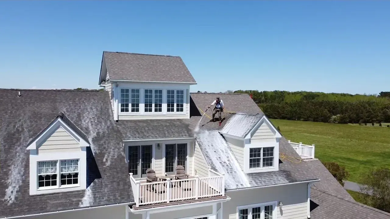 Une personne nettoie au nettoyeur haute pression le toit d'une grande maison par une journée ensoleillée.