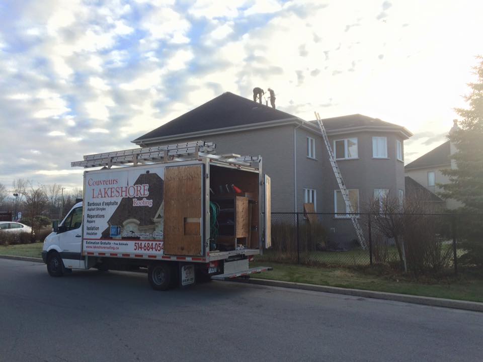 Une camionnette blanche garée devant une maison, des échelles menant au toit, des ouvriers sur le toit.
