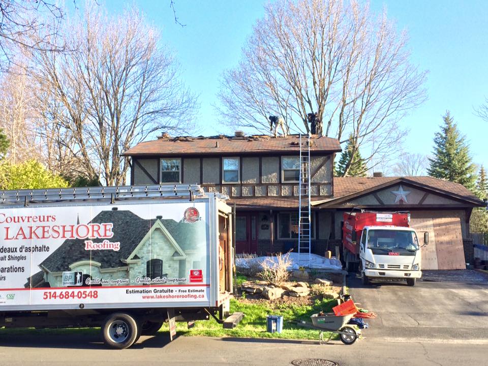 Des couvreurs travaillent sur le toit d'une maison, avec un camion de la société Lakeshore Roofing et un camion à ordures.