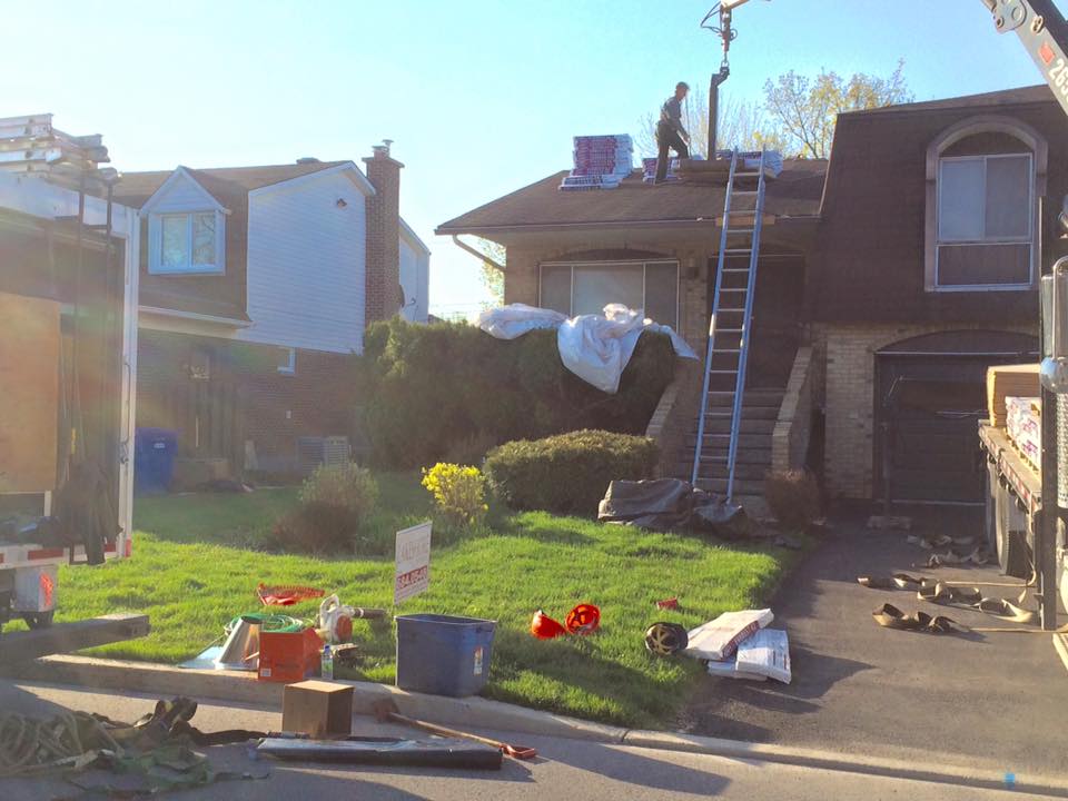 Une équipe de couvreurs travaille sur une maison à deux étages ; les matériaux sont éparpillés sur la pelouse.