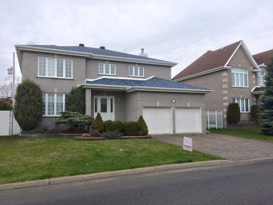 Maison de deux étages en briques grises avec des boiseries blanches, un garage double et une pelouse. Une autre maison est visible sur la droite.