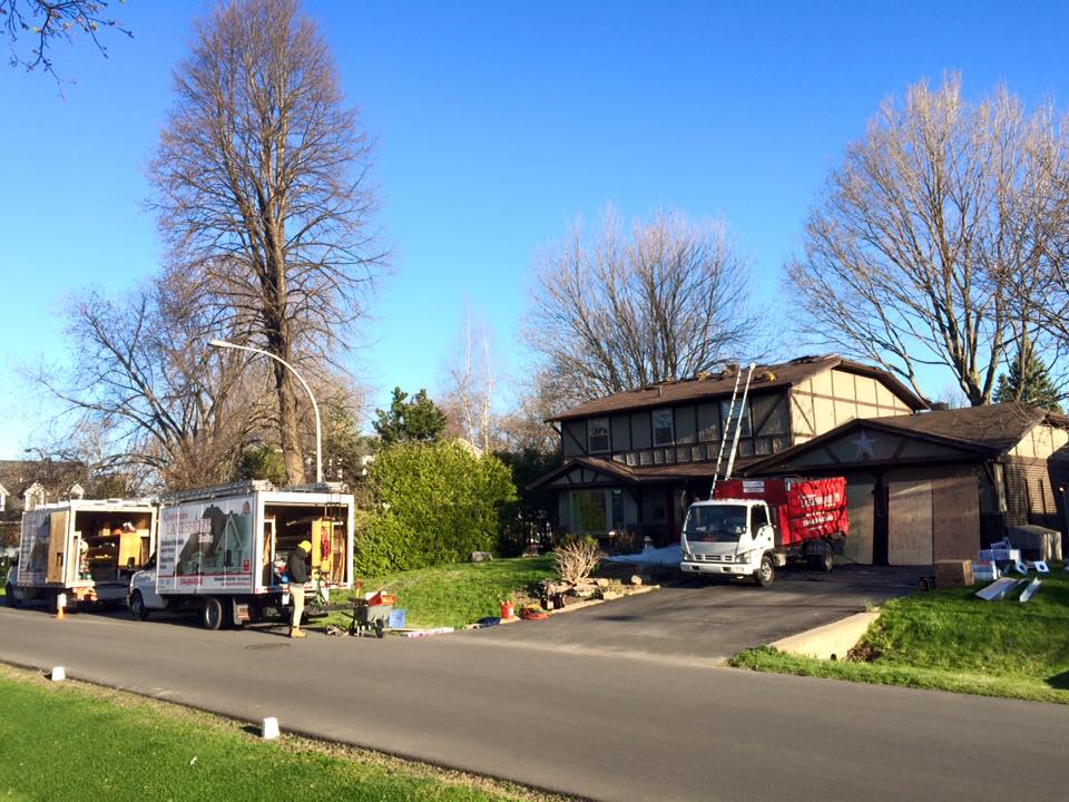 Des couvreurs travaillent sur une maison, des camions sont garés dans la rue. Toit marron, ciel bleu, arbres.