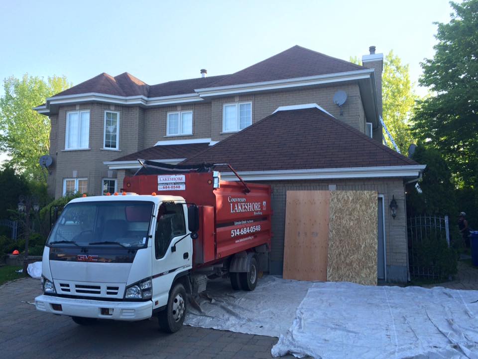Un camion-benne rouge est stationné devant une maison, probablement pour des travaux de toiture.
