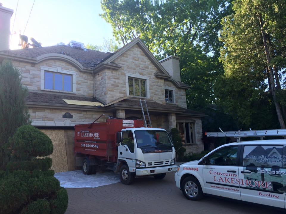 Des couvreurs travaillent sur une maison, un camion-benne et une camionnette sont garés dans l'allée.