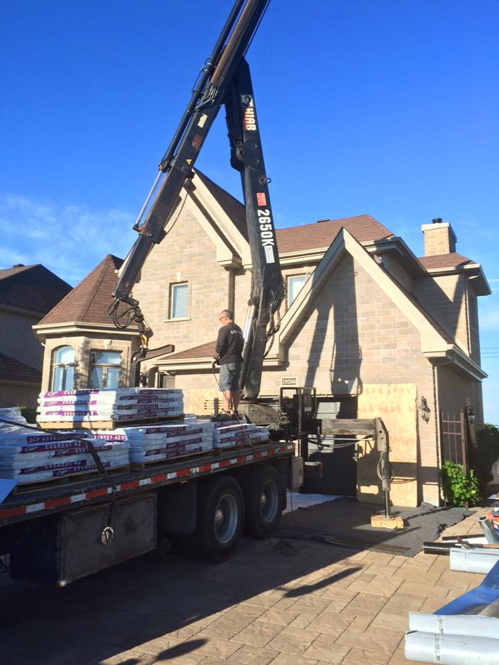 Une grue soulève des matériaux de toiture pour une maison à deux étages au toit brun et à l'extérieur en briques beiges, par une journée ensoleillée.