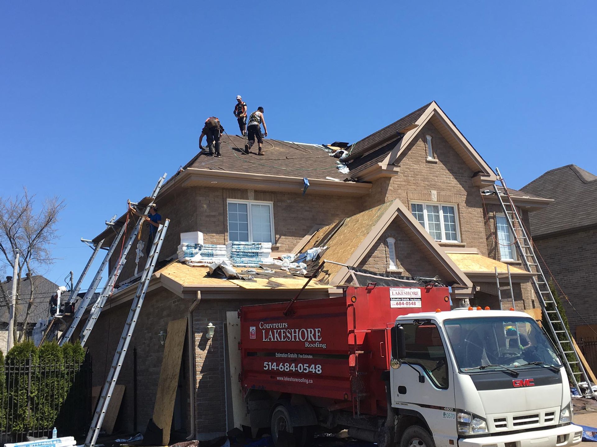 Une équipe de couvreurs travaille sur une maison en briques de deux étages, avec un camion-benne rouge garé devant, par une journée ensoleillée.