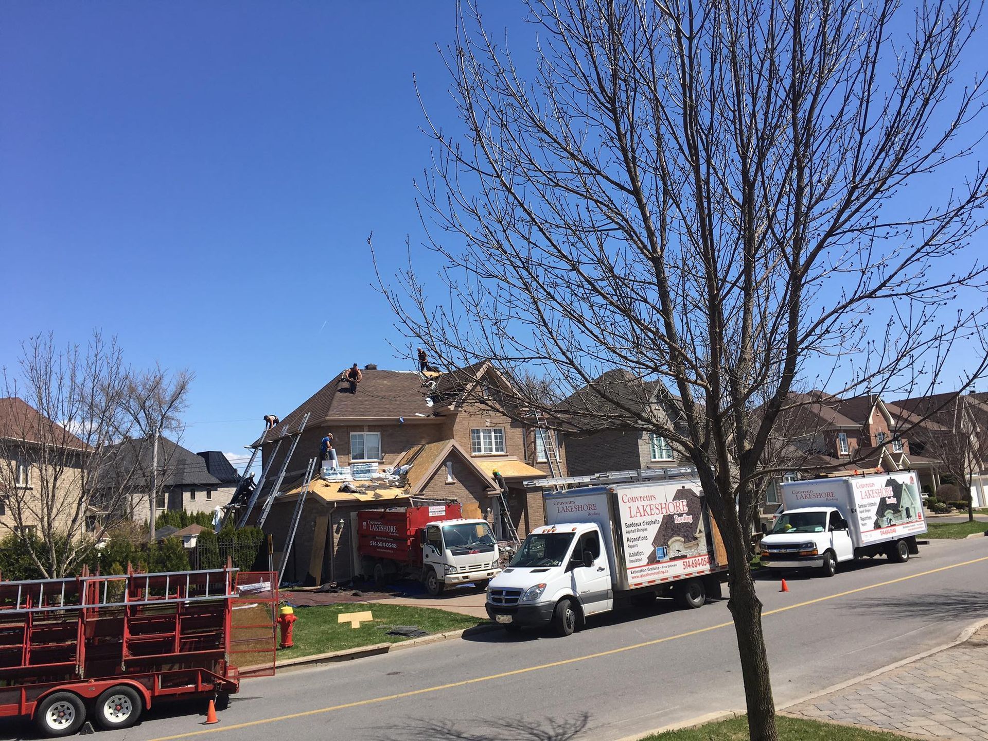 Des couvreurs travaillent sur une maison, des camions sont garés dans la rue sous un ciel bleu azur.