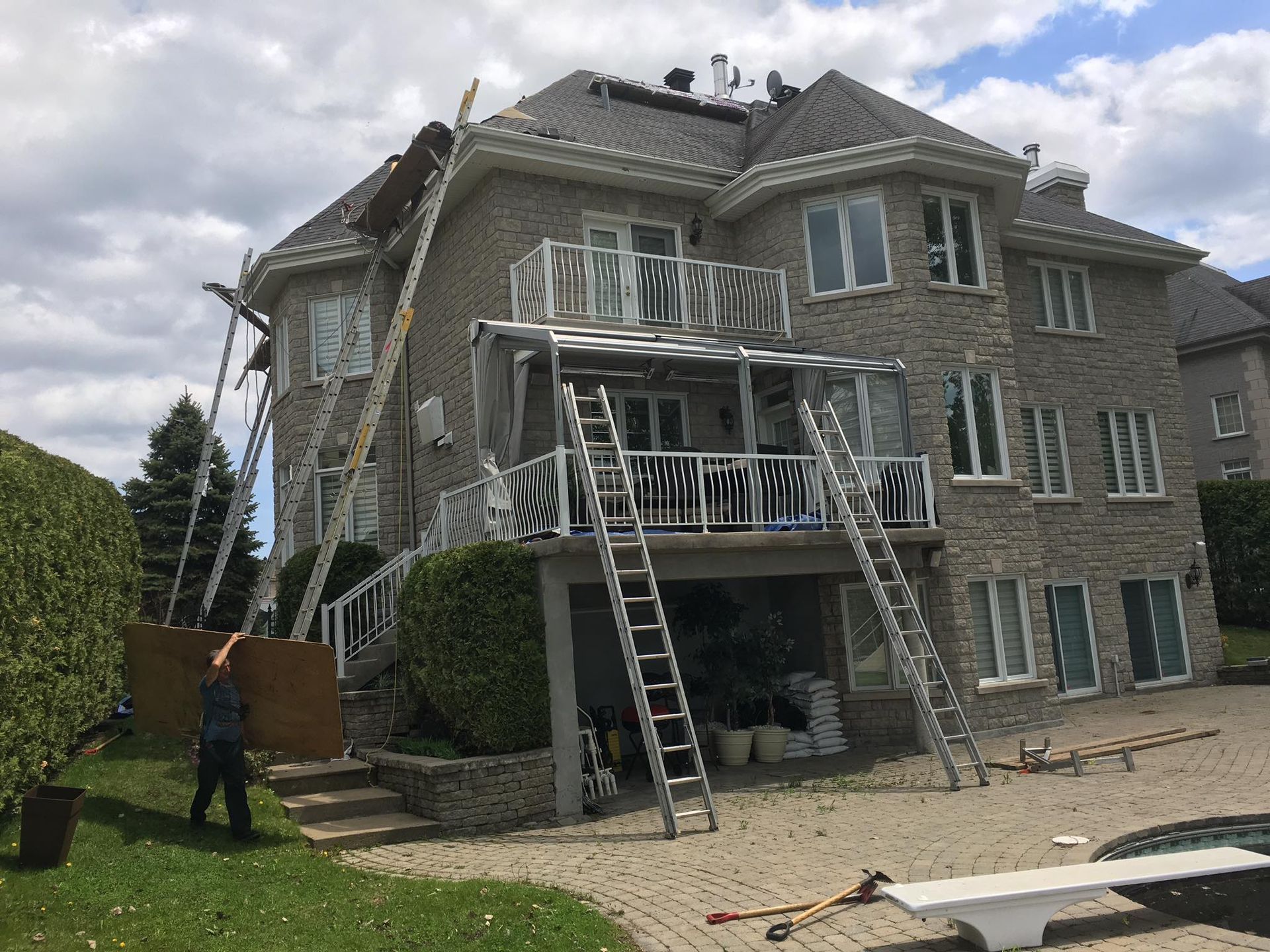 Des ouvriers et plusieurs échelles sur le toit d'une maison en pierre à plusieurs étages ; des matériaux sont en cours de déplacement.