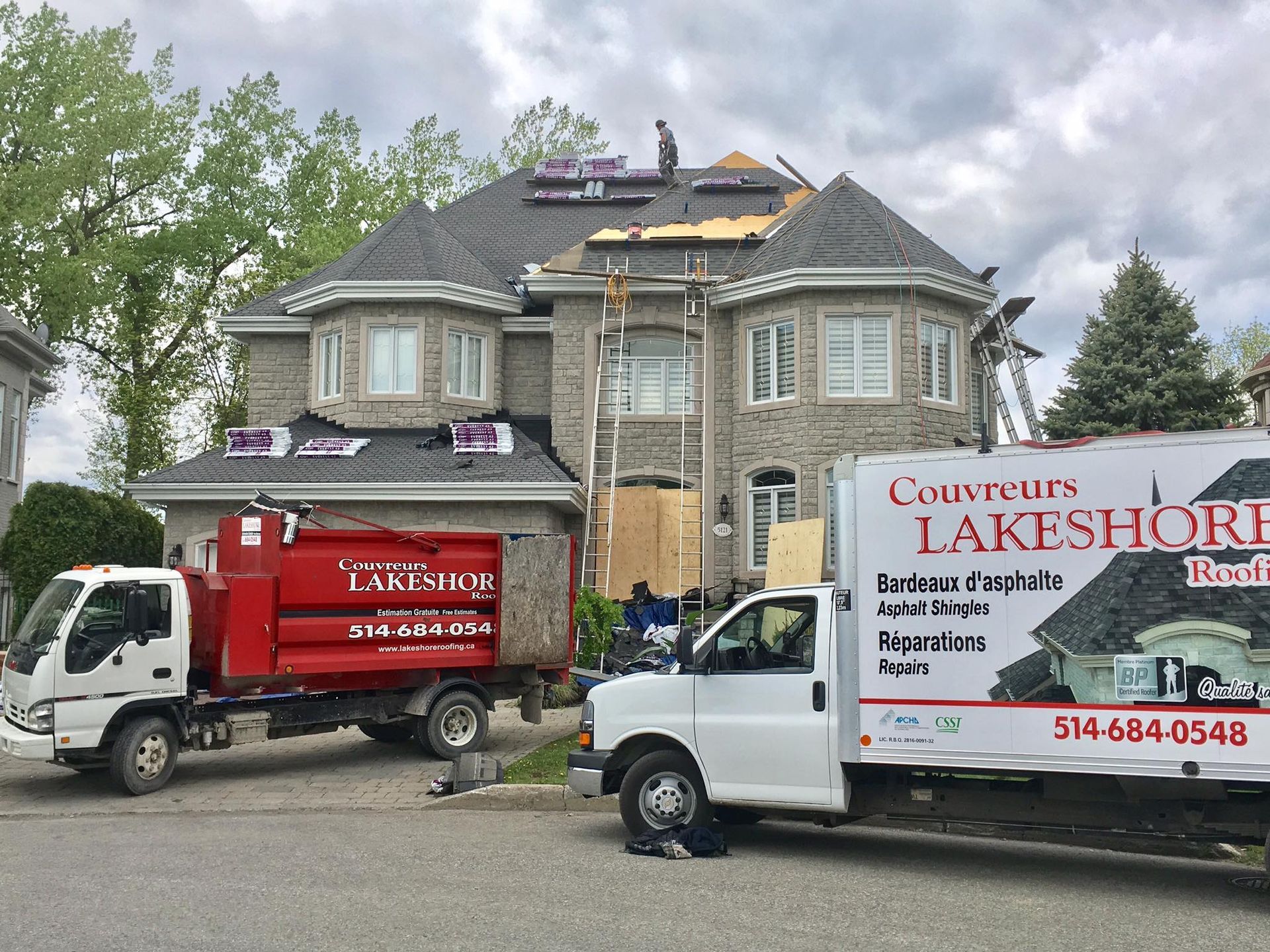 Des couvreurs travaillent sur une maison en pierre grise, deux camions sont garés dans l'allée.