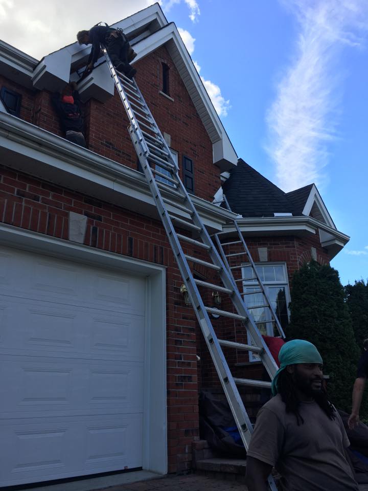 Des personnes travaillent sur des échelles sur le toit d'une maison en briques rouges ; une personne sourit au premier plan.