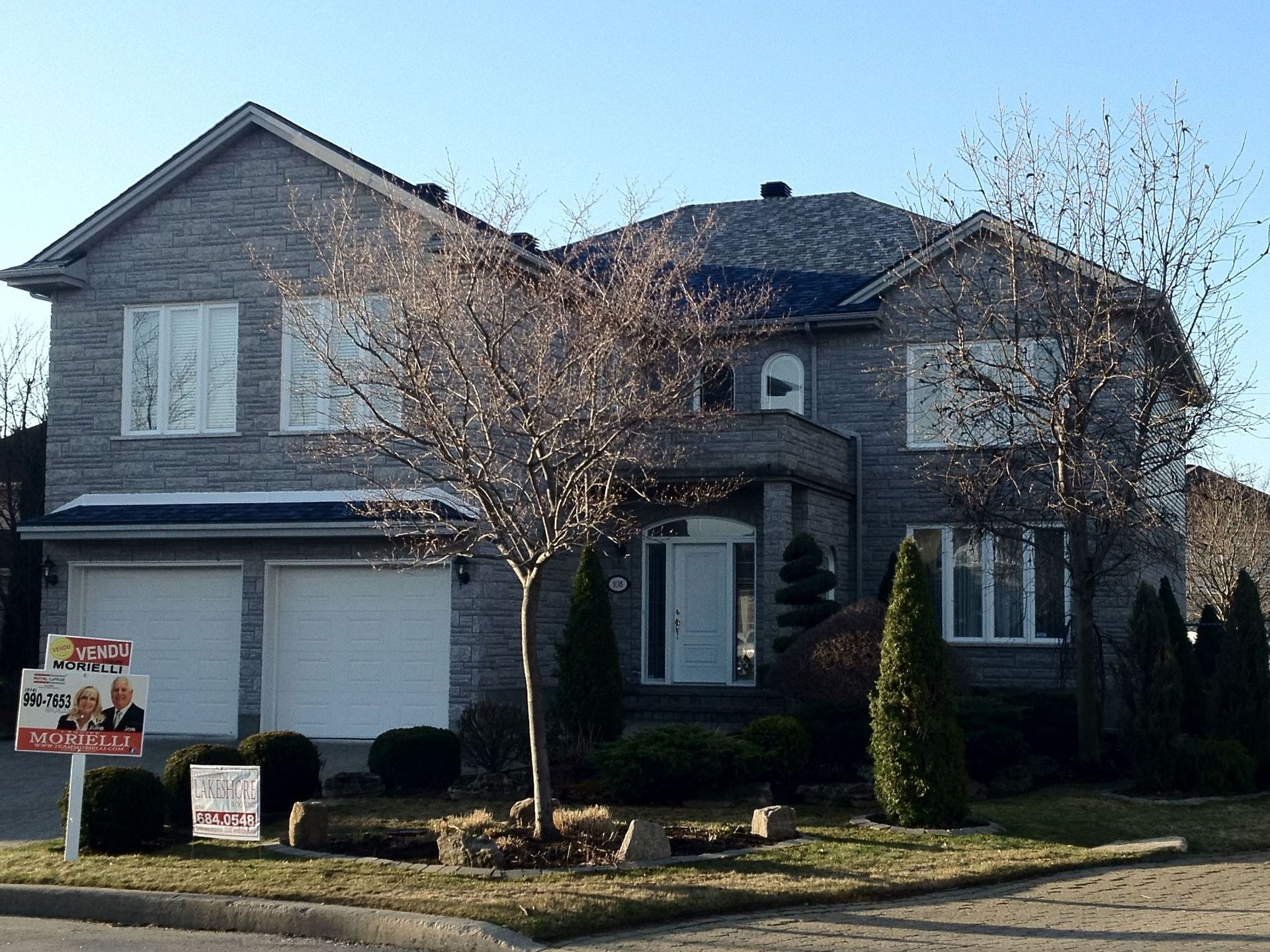 Maison en briques grises avec garage double, boiseries blanches et panneau « À vendre » par une journée ensoleillée.