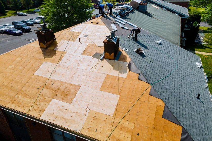 Toiture en cours de réparation par des ouvriers ; partiellement recouverte de bardeaux, charpente en bois apparente, avec cheminées sur un bâtiment en briques.