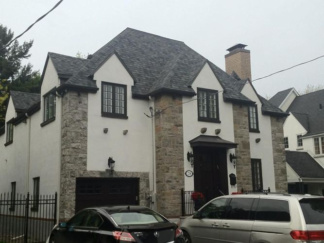 Maison à deux étages avec façade en pierre et stuc blanc, toit noir et garage. Voitures noires garées devant.