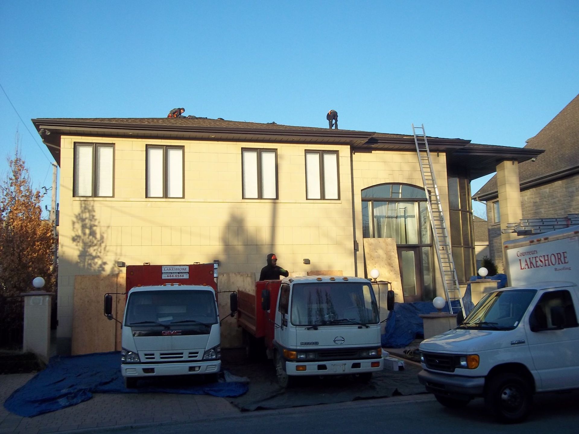 Maison avec camions stationnés, ouvriers sur le toit, échelle. Chantier, extérieur, journée ensoleillée.