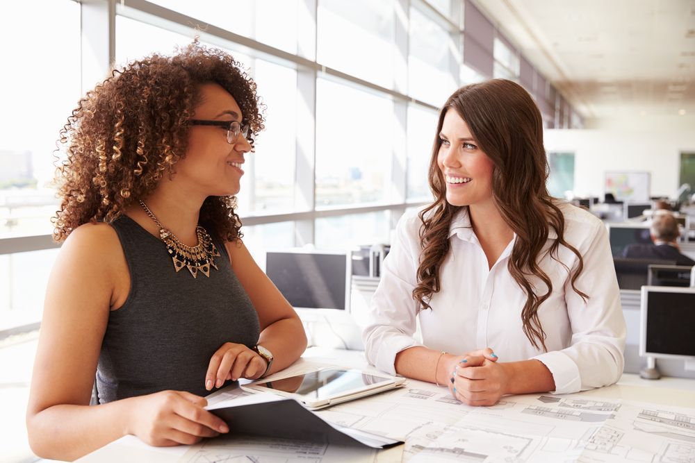 Deux femmes assises à une table dans un bureau lumineux, en pleine conversation, l'une tenant une tablette.