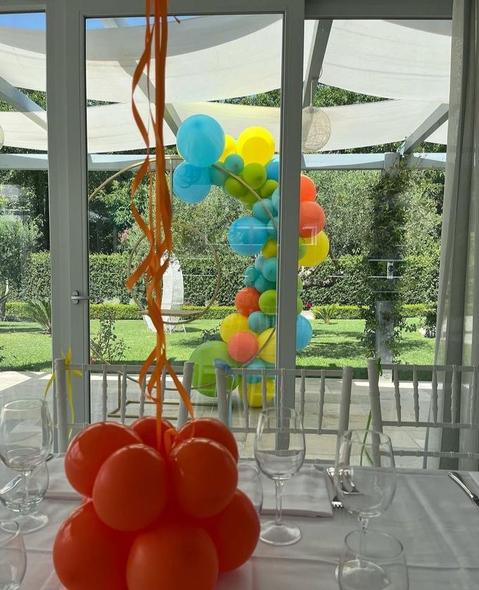 Composizioni di palloncini colorati su un patio di vetro, con palloncini arancioni in primo piano e un arco di palloncini blu-gialli sullo sfondo.