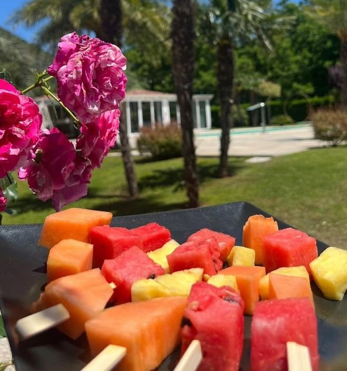 Cubetti di anguria, papaya e ananas su un piatto nero all'aperto, con fiori rosa e palme sullo sfondo.