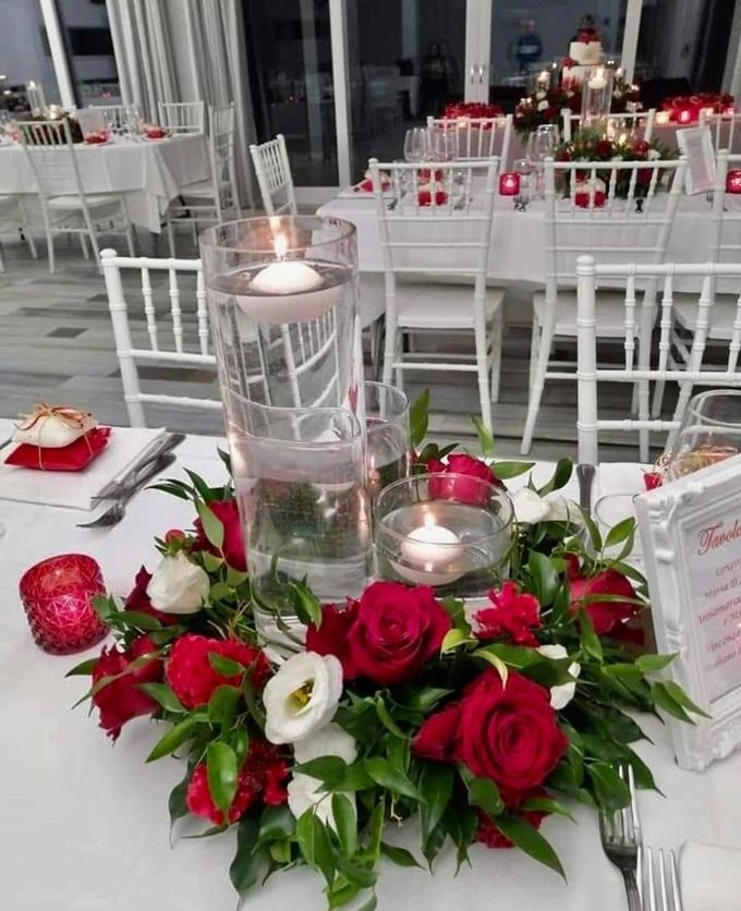 Centrotavola bianco per banchetti con rose rosse, verde e candele accese in un vaso trasparente.
