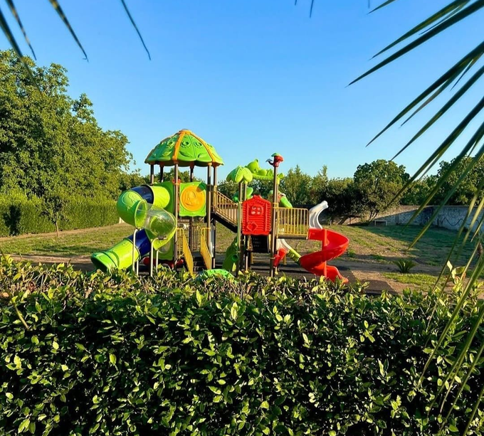 Un coloratissimo parco giochi con scivoli e strutture per l'arrampicata in un parco soleggiato, visibile dietro una siepe.
