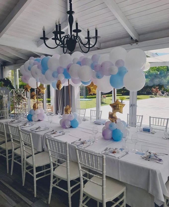 Un tavolo da banchetto bianco all'aperto, sotto un arco di palloncini color pastello e un lampadario nero, su un patio coperto.