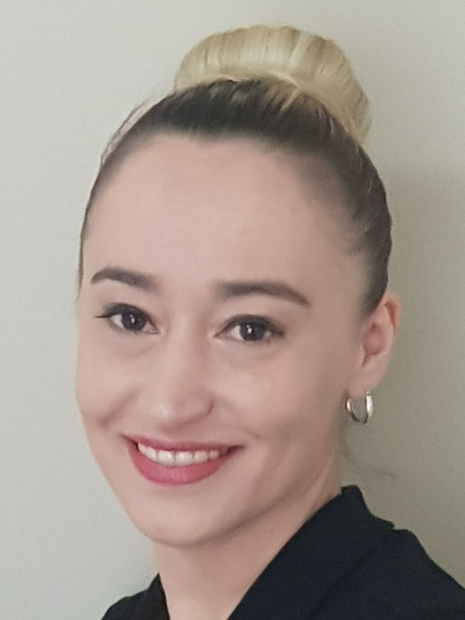 Smiling person with blonde hair in a bun, wearing hoop earrings and a black top against a plain wall
