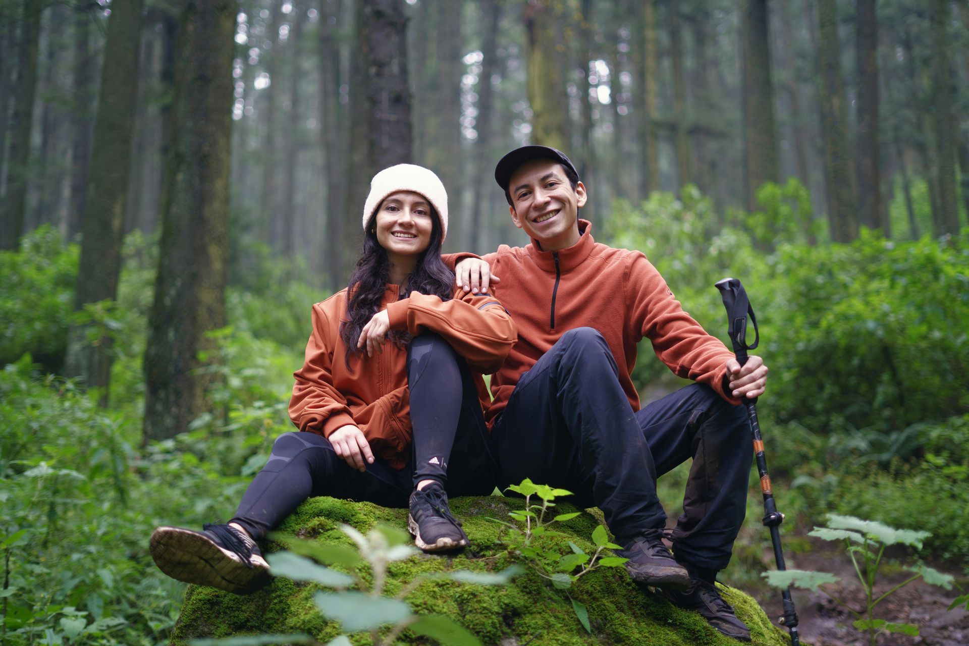 Two hikers in orange jackets sit smiling on moss in a misty forest, holding trekking poles.