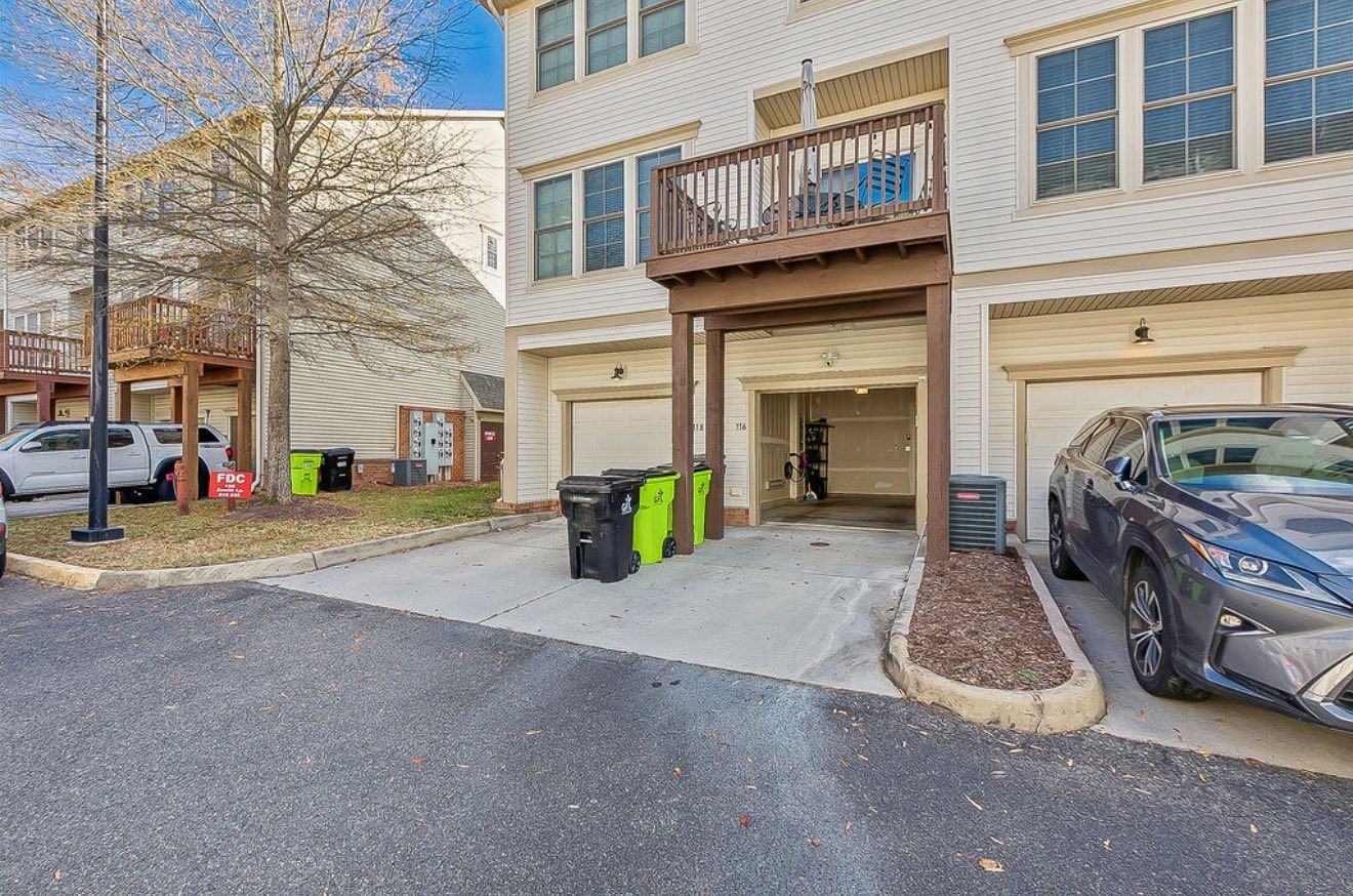 Apartment building parking area with a balcony, garage entrances, trash bins, and parked cars