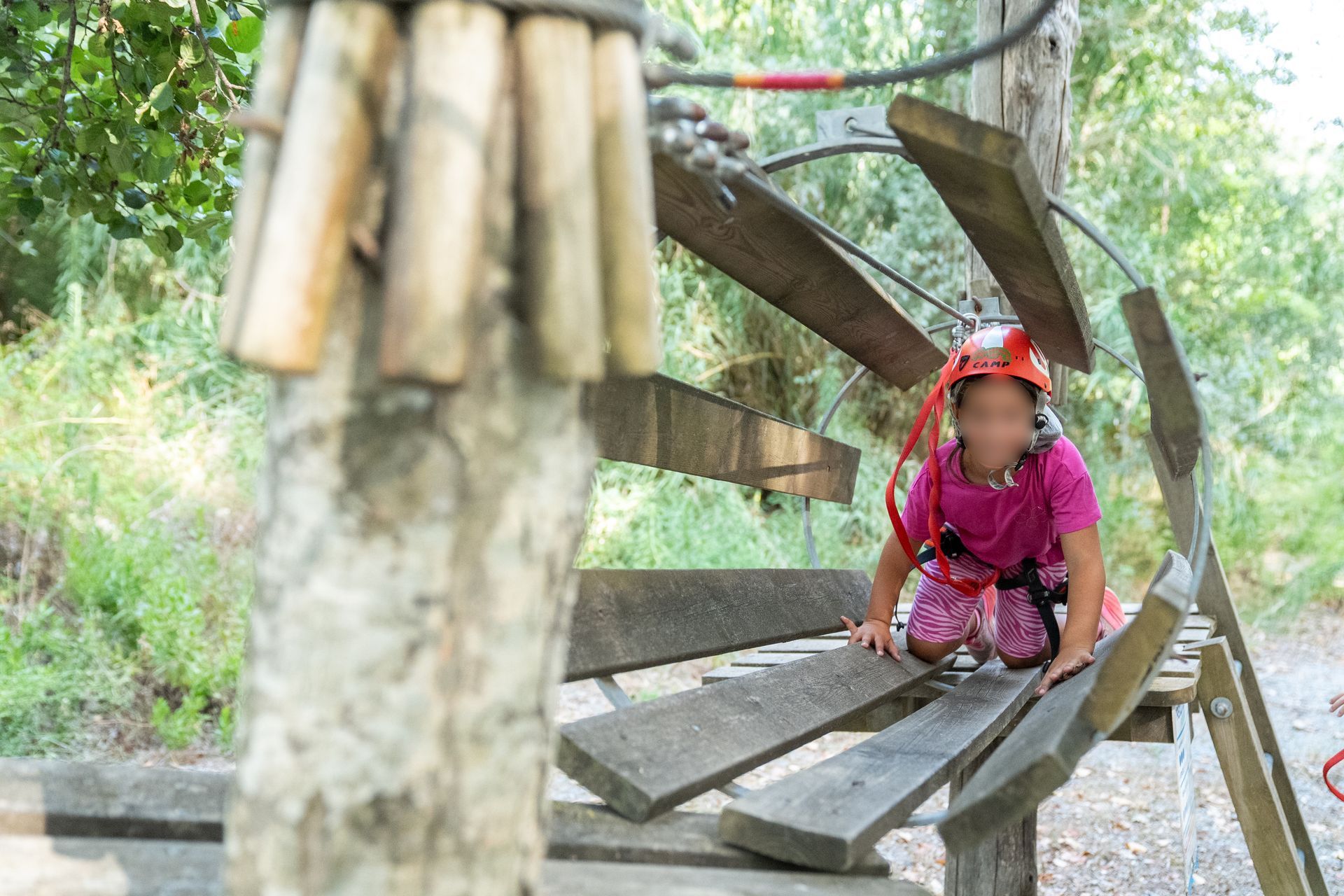 Una persona con una maglietta rosa è accovacciata su una struttura di legno in un ambiente esterno verde.