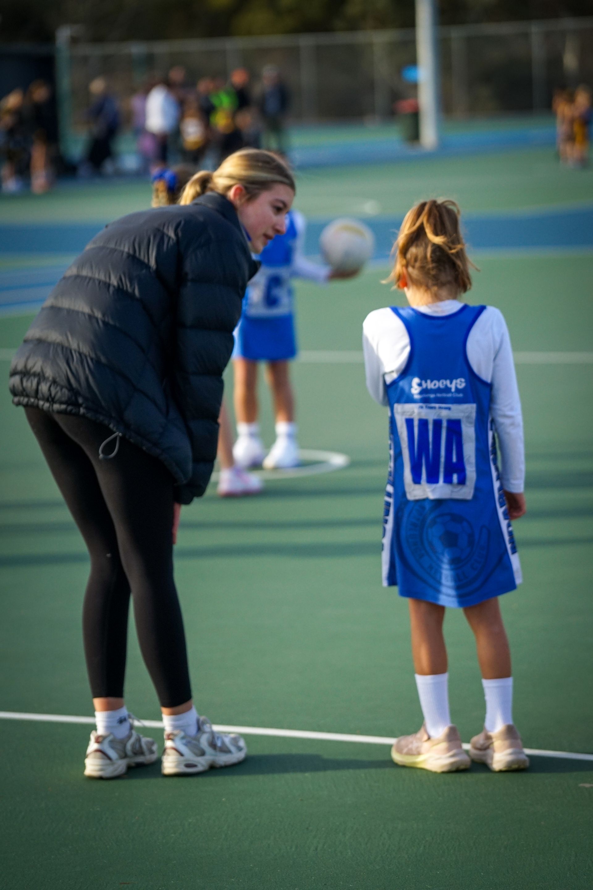 Woman in black coat coaches girl in blue netball uniform on a green court.