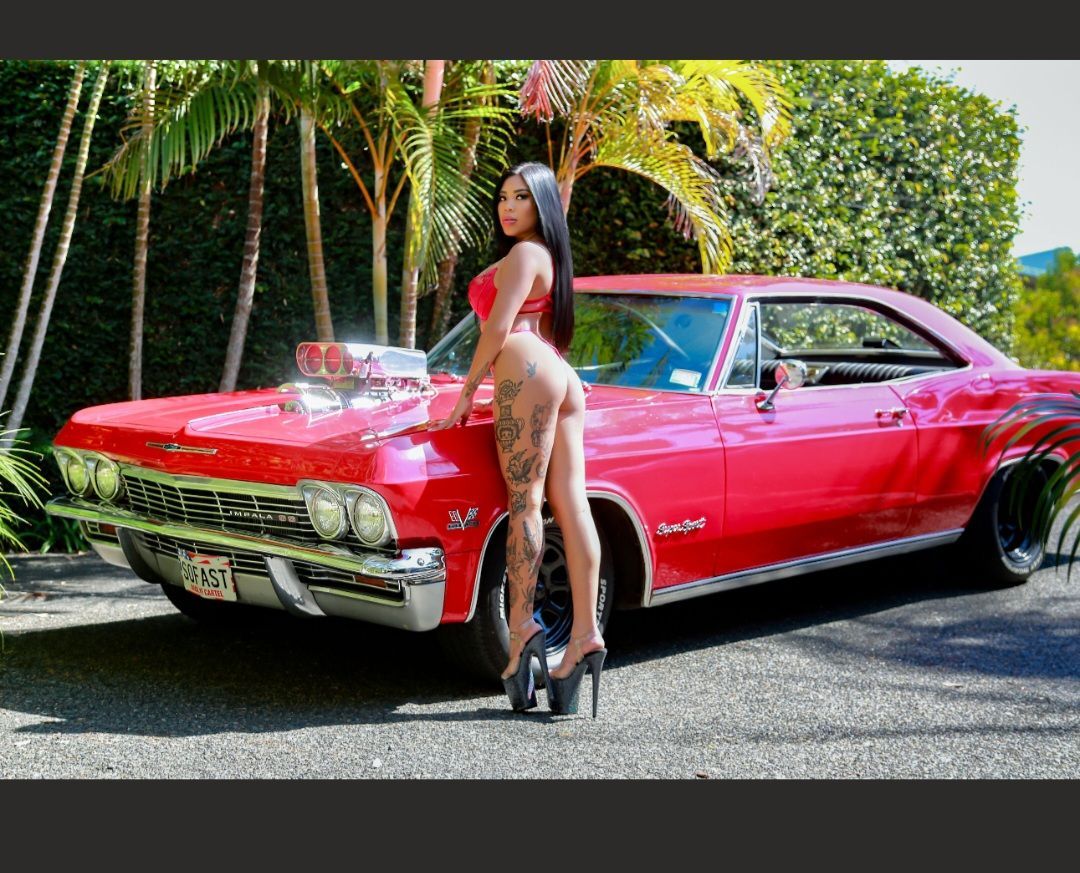 Woman posing beside a red classic car on a palm-lined driveway, wearing a red top and high heels