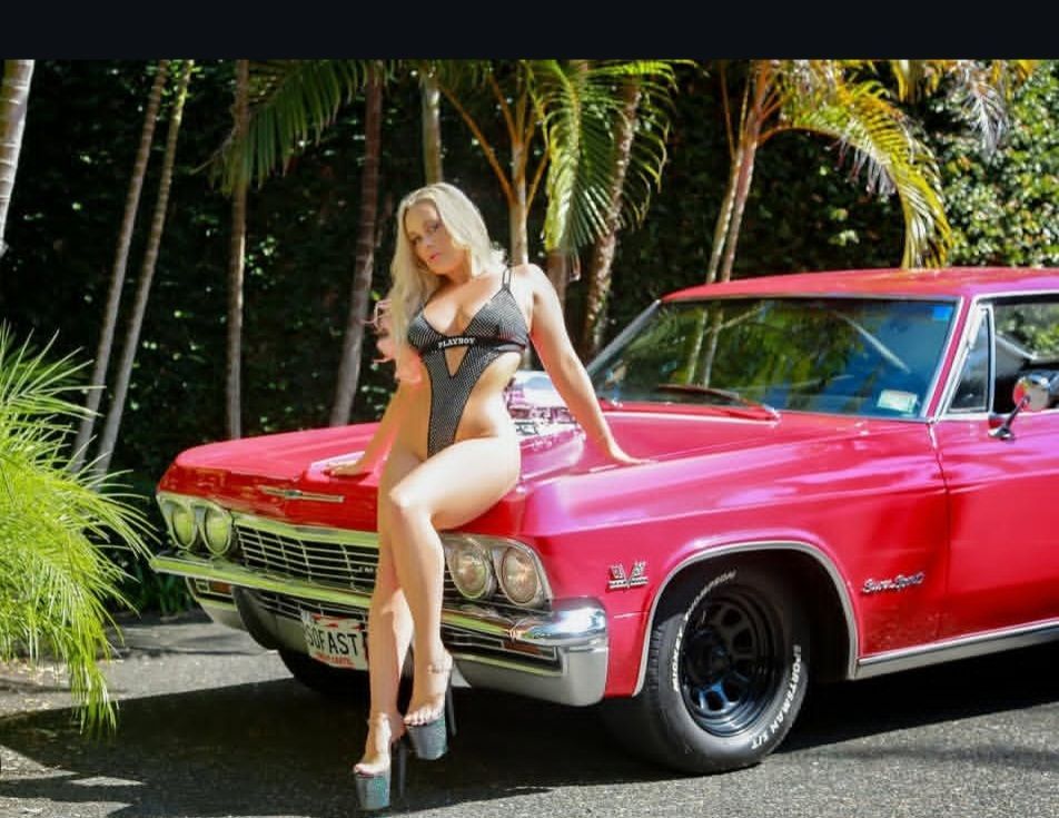 Woman in a black swimsuit posing on a bright pink classic car in a tropical setting