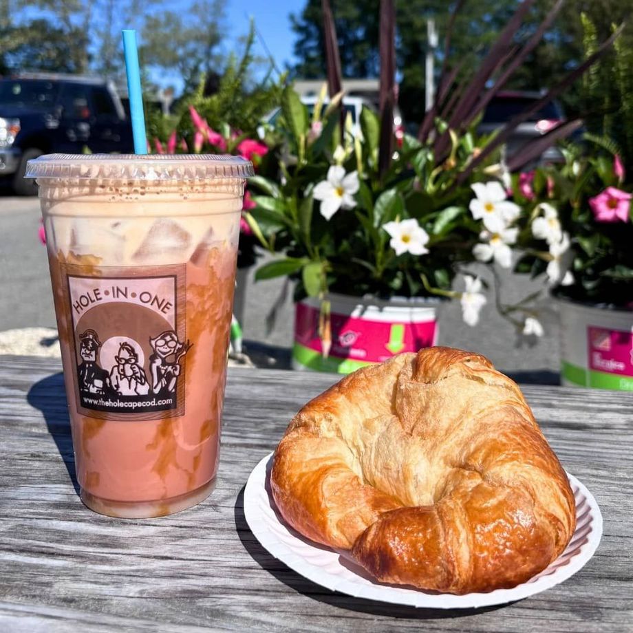 Iced coffee with a straw and croissant on a plate, set on a wooden table outdoors with flowers.