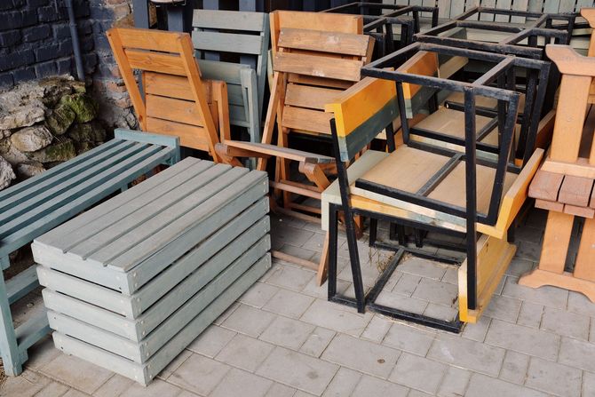 Stacked wooden pallets and metal frames on a paved outdoor surface against a stone wall.