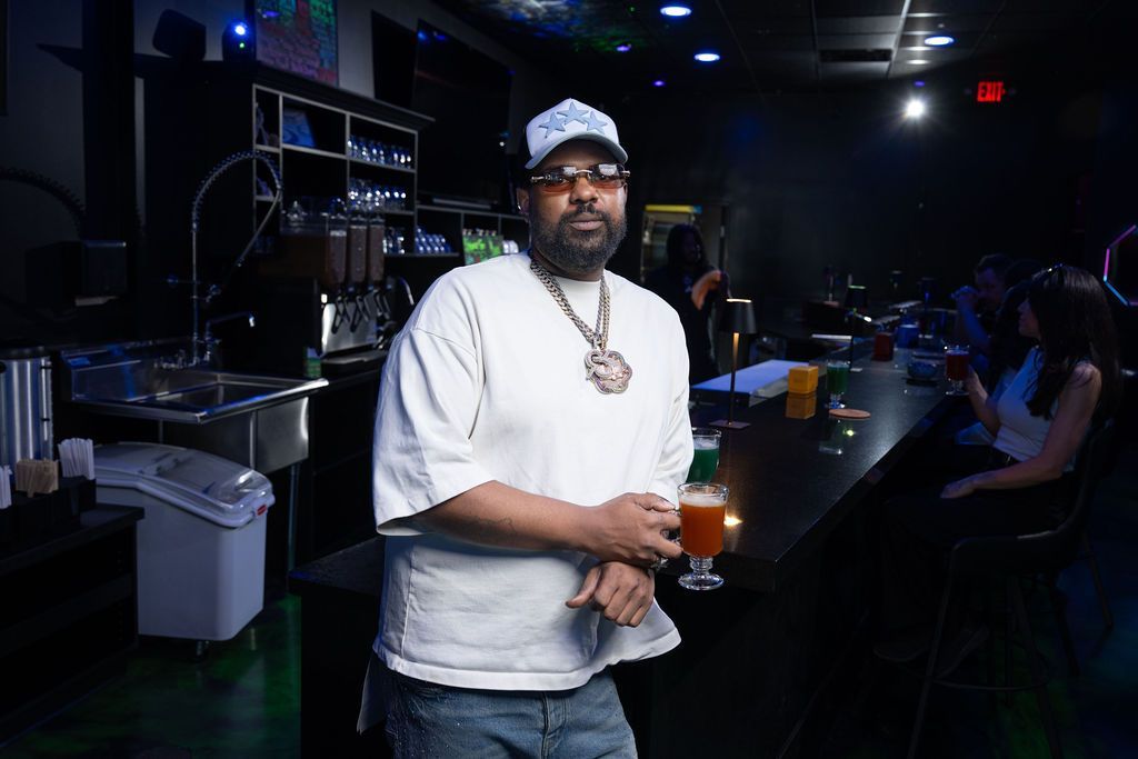 Man standing at a dim bar counter holding a drink, with seated patrons in the background