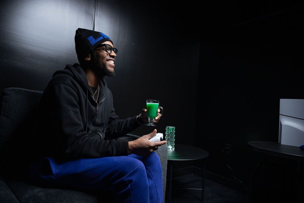 Person seated in a dim lounge, smiling and holding a green drink beside a small table.