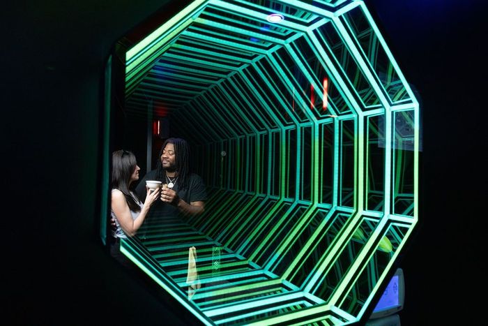Two people in a neon-lit tunnel with geometric light frames, facing each other and talking
