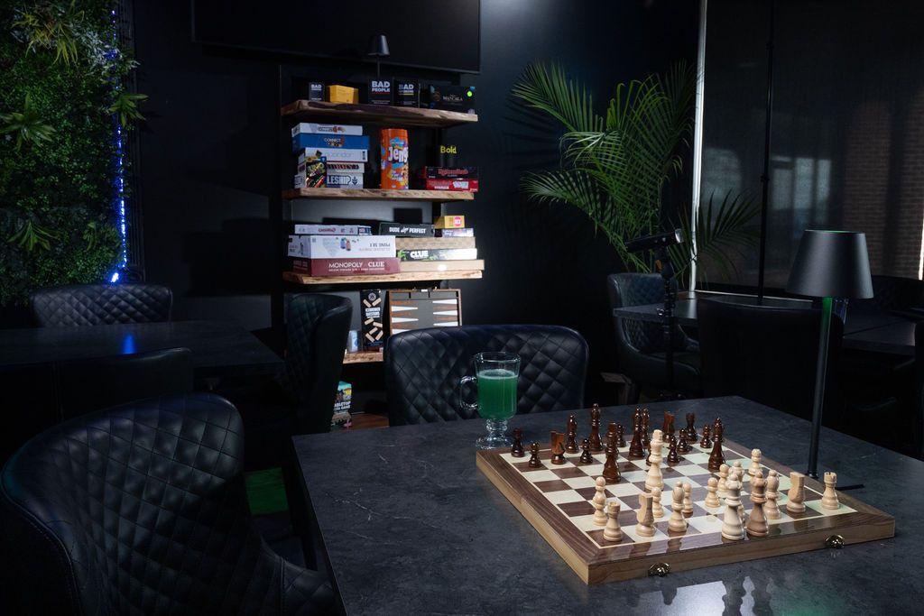 Dim lounge with a chessboard on a table, surrounded by dark chairs, plants, and a bookshelf in the background.