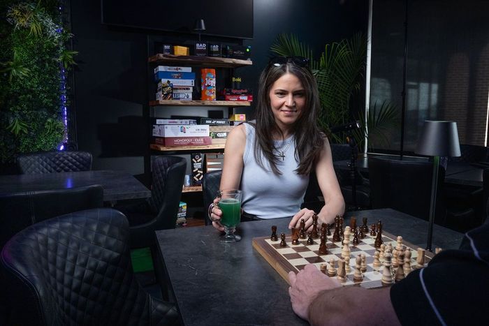 Woman playing chess in a dimly lit lounge, smiling across a board with drinks nearby