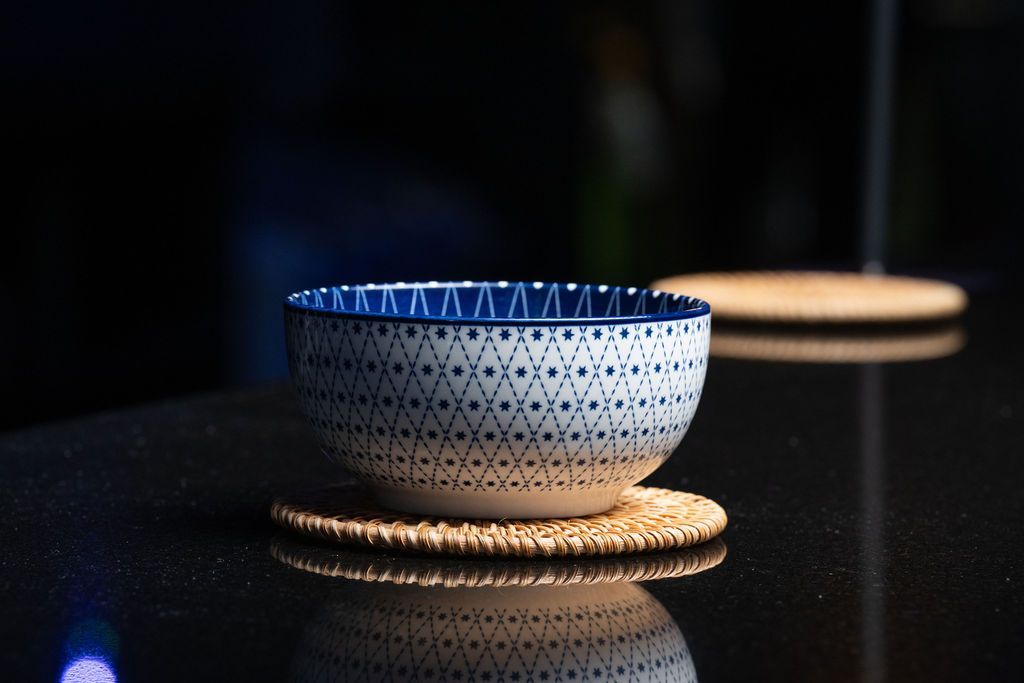 Blue-and-white patterned bowl on a woven coaster, with its reflection on a glossy black surface