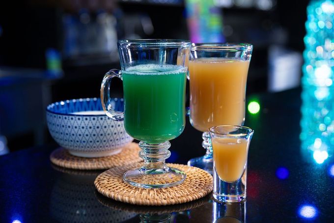 Two colorful cocktails on coasters, one green and one orange, on a glossy bar counter