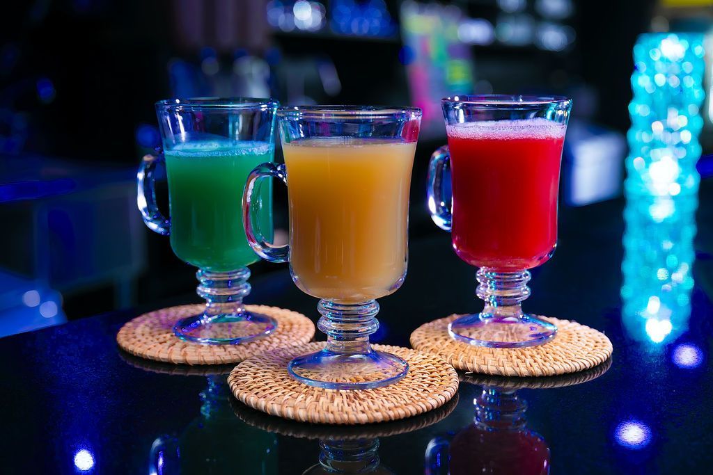 Three colorful cocktails in clear mugs on coasters at a dimly lit bar countertop