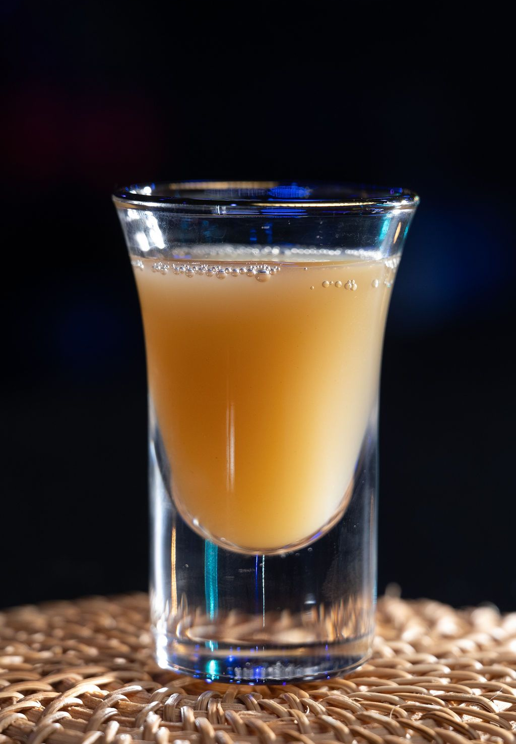 A shot glass of cloudy beige drink on a woven mat against a dark background.