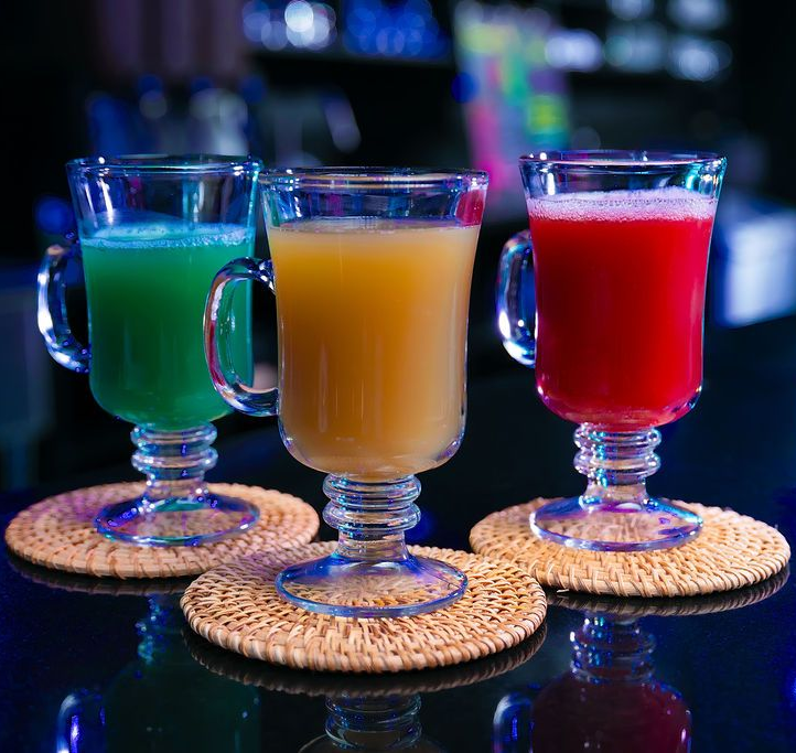 Three colorful cocktails in stemmed glasses on woven coasters at a dimly lit bar
