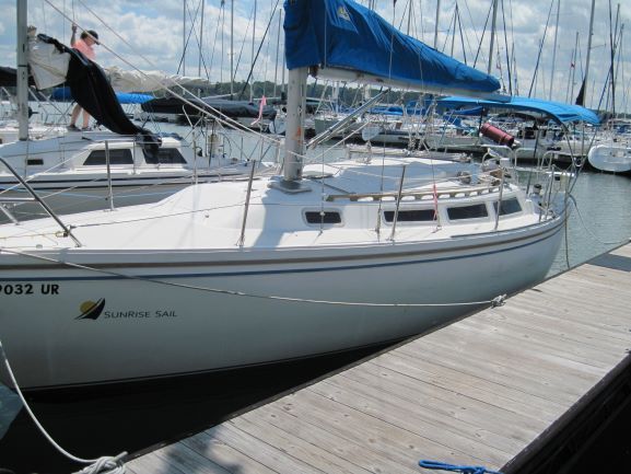 White sailboat at a dock, blue sail up. Sunny day, other boats in background.