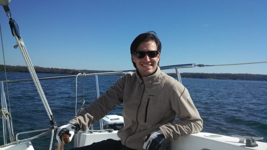 Man smiling, steering a sailboat on a sunny day. Blue sky and water, with land in the background.